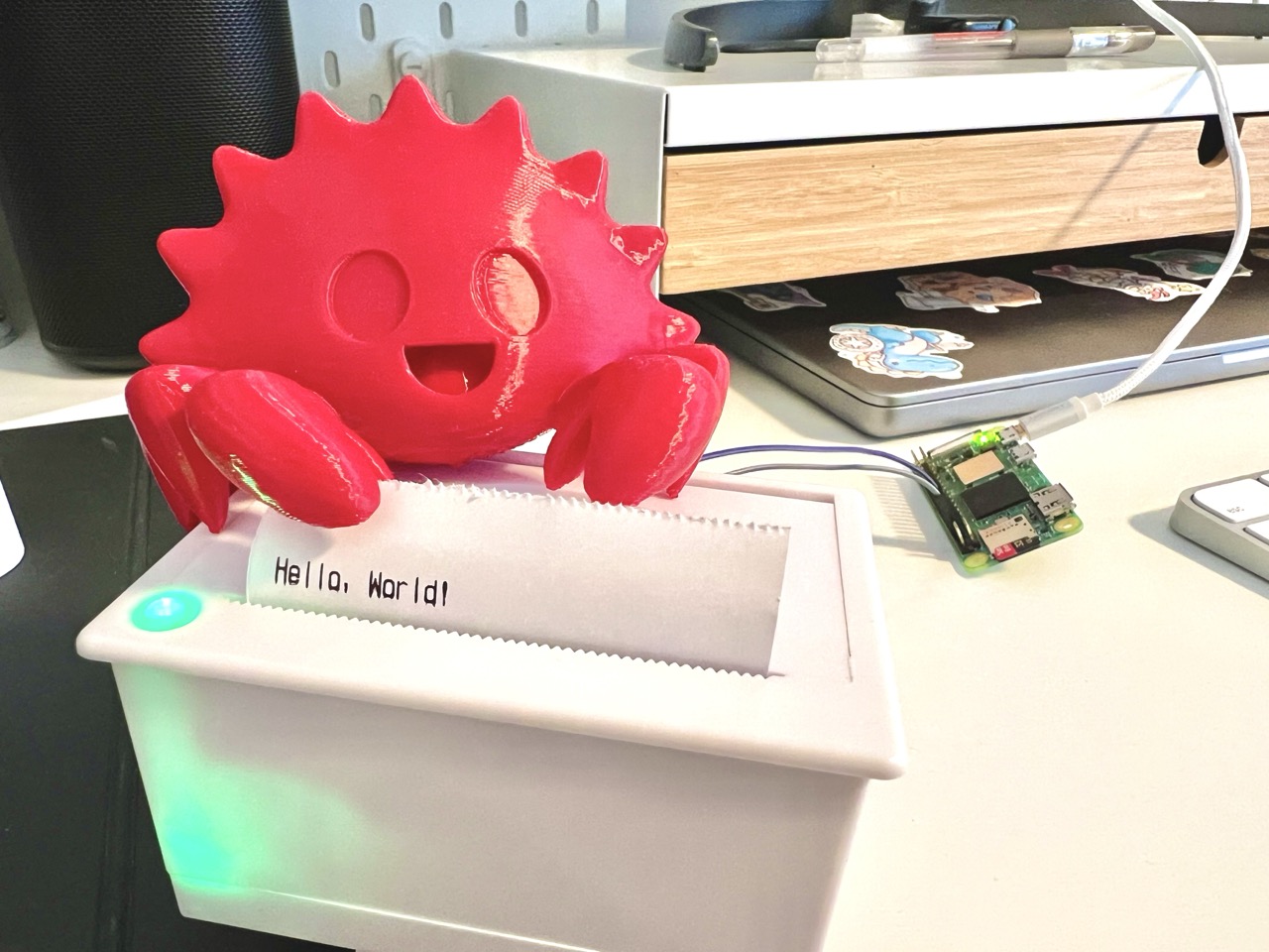 Thermal printer with a piece of paper coming out with the text 'Hello World'. On top of the printer is a 3D-printed crab, the mascot of the Rust programming language. In the background, wires connect the printer to a Raspberry Pi Zero 2 W.