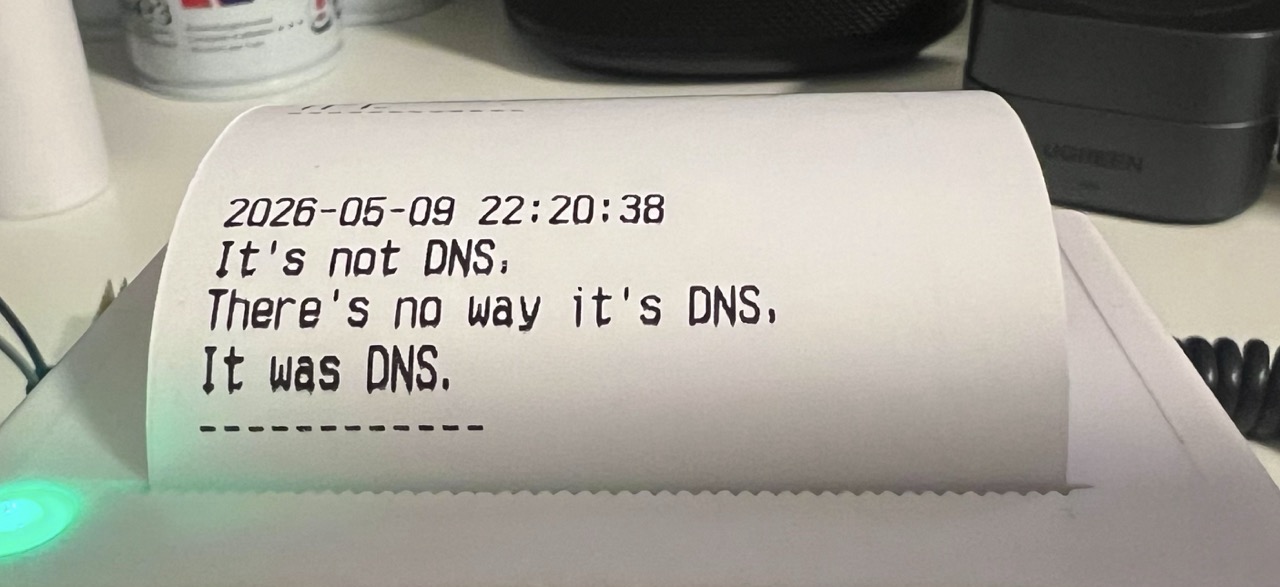 A small thermal printer with a piece of paper hanging out of it, reading: "2026-05-09 22:20:38, It's not DNS, There's no way it's DNS, It was DNS."